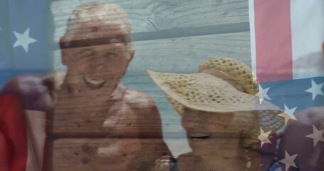 Smiling group of seniors in swimwear posing at beach shoreline, with straw sun hat, US flag