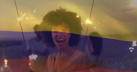 Laughing woman in loose striped top holding sparklers at beach sunset, alongside friend in T-shirt
