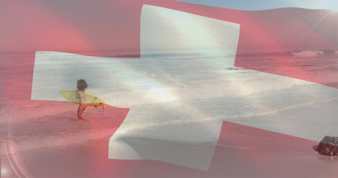 Standing woman surfer holding yellow surfboard in shallow surf at sandy beach, Swiss cross overlay