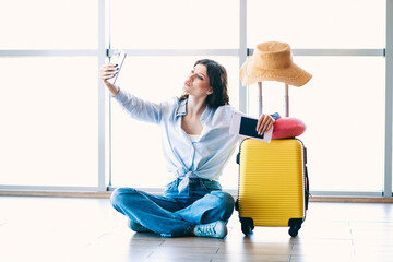 Charming Young woman sitting on the floor with travel items in an airport terminal taking a selfie