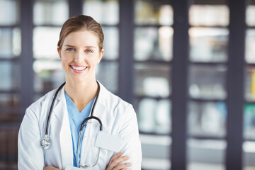 Female physician standing confidently with arms crossed in clinic corridor wearing stethoscope