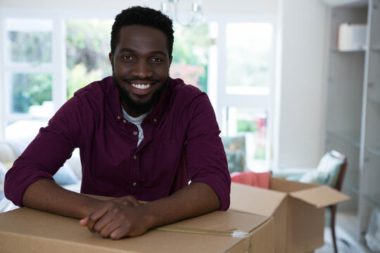 African American man leaning on cardboard box under chandelier at home surrounded by moving boxes