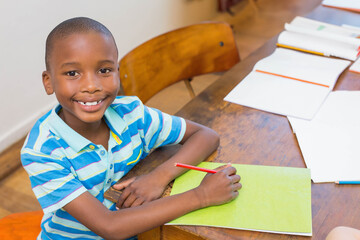 African American boy student sitting at desk in classroom holding red pencil over green notebook