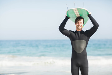 Man wearing black wetsuit holding green surfboard overhead on shore under clear sky, copy space
