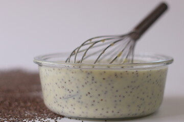 Chia custard in a glass bowl with whisk, surrounded by chia seeds on a white surface