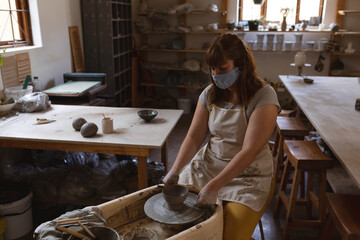 Female potter shaping wet clay bowl on spinning wheel in ceramics workshop, with tools on tables