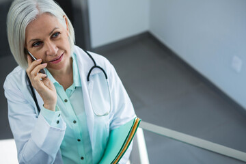 Woman with lab coat and stethoscope in corridor speaking on smartphone, holding files, copy space