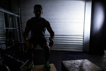 Athlete squatting on wooden plyo box in dimly lit gym with squat rack and punching bag