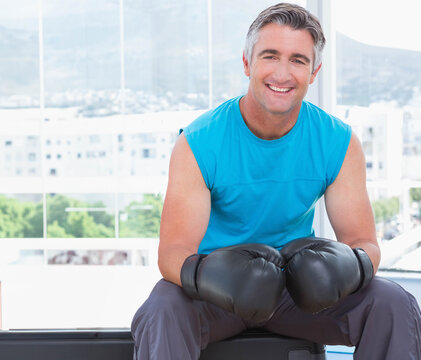 Middle-aged man sitting on bench by glass windows in gym wearing turquoise top and boxing gloves - Powered by Adobe