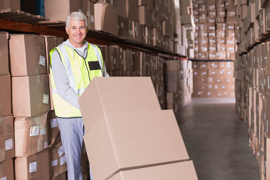Senior male warehouse worker wearing neon vest pushing dolly with cardboard boxes down rack aisle