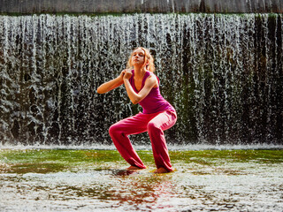Young fitness girl model is standing in river water with stunning waterfall background. Girl is...