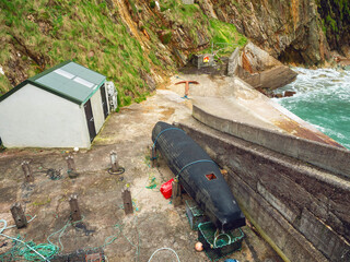 A black boat is sitting on the beach next to a shed. The boat is covered in a tarp and is...