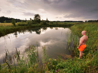 A man is jumping into a pond wearing an orange swimsuit at dusk. The sky is cloudy and the water is...