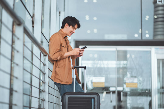 Happy Asian man using mobile smartphone standing with suitcase booking travel tickets via mobile application at airport terminal, Browse Internet, flight check in, Tourist journey trip concept