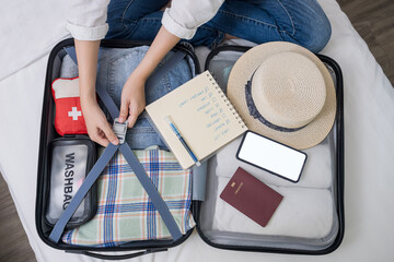 top view of woman hand packing suitcase for trip, preparing for summer holidays abroad with travel medicine first aid kit, cloth, accessory, phone mockup on open luggage, notebook for checklist item