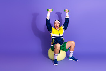 Energetic young man in retro sportswear lifting dumbbells while sitting on a fitness ball against a...