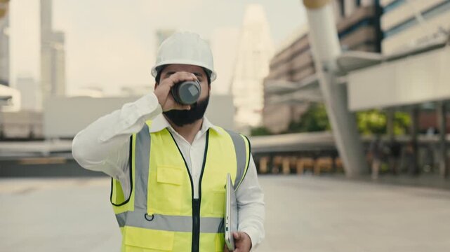 Portrait of Indian engineer holding coffee and laptop under arm walking across city square. Friendly technician with drink and computer smiles ready for shift at construction site