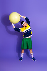 Young athletic man in retro sportswear with exercise ball posing against vibrant purple background...