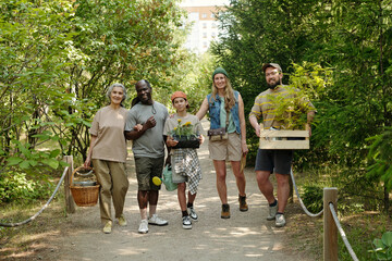 Group of diverse adults and one teenager walking outdoors on forest path carrying potted plants and gardening supplies, smiling and looking forward, surrounded by green trees