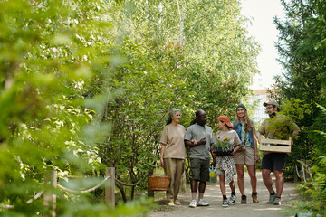 Group of diverse young adults and one senior Caucasian woman walking outdoors on forest path carrying plants and gardening supplies, engaging in conversation and smiling together