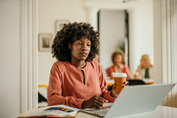 Focused black businesswoman working on laptop with coffee in hand at modern office
