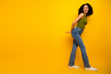 Cheerful young woman posing casually in green top and jeans against a vibrant yellow background...