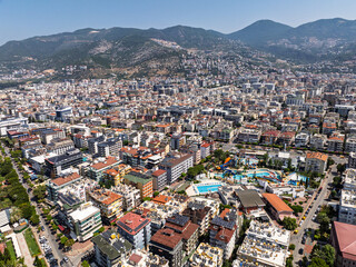 Obraz premium Aerial view of Alanya city in Turkey. Dense urban buildings, Mediterranean coastline, green hills and mountains, from drone during clear summer day. Aerial view of buildings and hotels in Alanya city