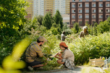 Caucasian man helping Caucasian girl planting sapling in urban garden while senior Caucasian woman...