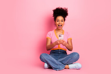 Young woman with joyful expression holding smartphone seated on pink background, showcasing vibrant...