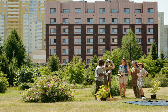 Group of diverse young adults and teenagers standing together in urban park, holding gardening tools and plants, engaging in community gardening activity with residential buildings behind - Powered by Adobe
