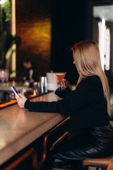 Woman Relaxing at Stylish Indoor Bar Holding Cocktail and Using Smartphone