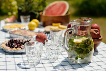 Glass pitcher with lemon slices and mint standing on checkered tablecloth near empty glasses, fresh apples, watermelon, berry pie, and lemon outdoors during summer picnic