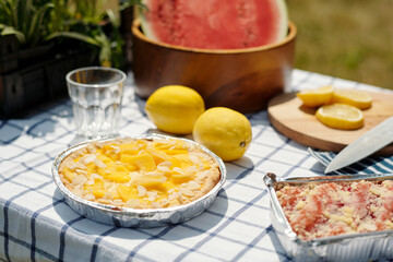 Freshly baked peach tart with almond slices and fruit crumble dessert sitting on checkered tablecloth near lemons, glass of water, and sliced watermelon outdoors