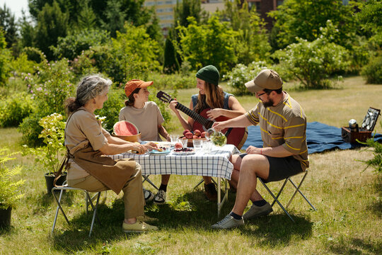Caucasian senior woman, Caucasian teenager boy, Caucasian teenager girl, Caucasian young adult man sitting outdoors at picnic table, teenagers playing guitar, group sharing food