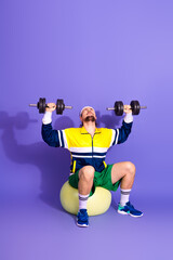 Young male athlete lifting dumbbells in a retro sports outfit while sitting on a stability ball...