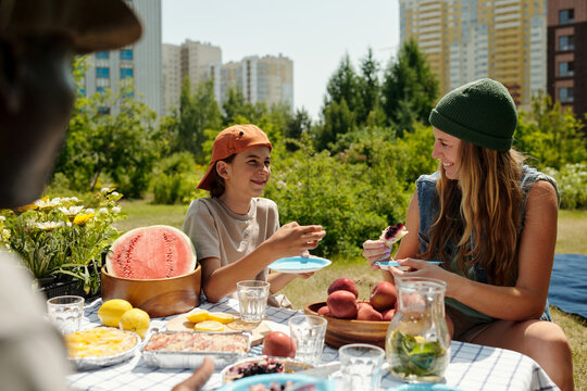 Caucasian teenage girl and Caucasian teenage boy sitting outdoors at picnic table smiling and eating fruit together with Black man