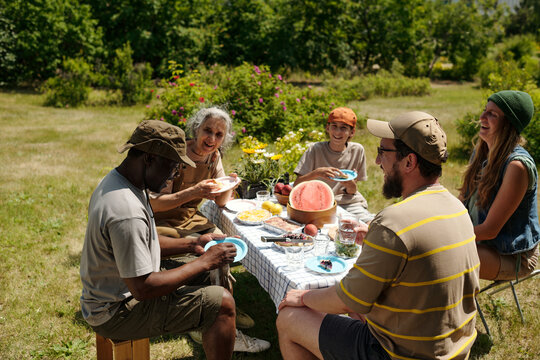Group of multiethnic adults and teenagers sitting outdoors around picnic table sharing food and laughing, senior Caucasian woman smiling, Black man holding plate, summer garden setting