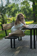 A young girl in a white dress sits alone on a wooden bench surrounded by lush greenery, her posture and distant gaze quietly conveying a deep sense of sadness and solitude.