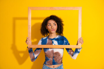 Portrait of a stylish young woman holding a wooden frame against a vibrant yellow background with a...