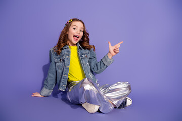 Smiling young girl in a denim jacket and shiny pants posing against a vibrant purple background...