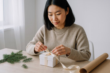 asian woman wrapping gift box with ribbon at home, creating festive atmosphere in warm natural light during holiday season