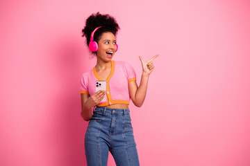 Cheerful young woman with headphones and smartphone posing on pink background showing energetic...