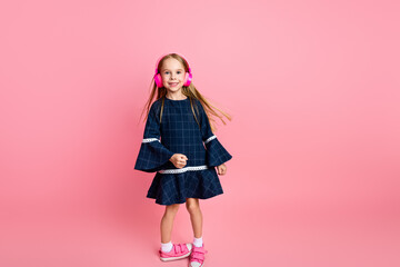 Smiling young girl in stylish outfit with headphones on pink background enjoying music