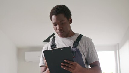 African American young male worker holding clipboard and reading task list in bright apartment. Technician in overalls focusing on renovation details indoors near air conditioner and lamp.