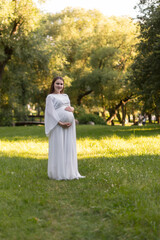 A portrait of a beautiful pregnant woman in a long white maternity dress, on a sunny summer day in...