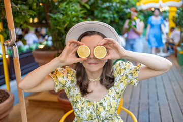 A woman in a floral dress playfully holds lemon halves in front of her eyes, creating a fun and fresh atmosphere in a cozy outdoor setting