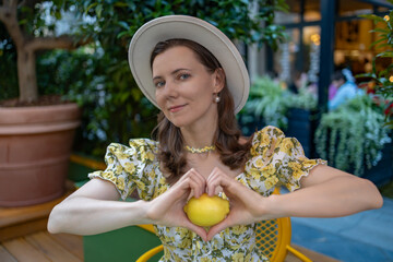 A woman in a yellow floral dress and a hat sits outdoors, holding a lemon in the shape of a heart, surrounded by greenery