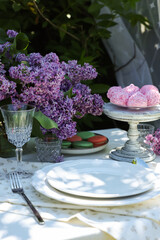 Beautiful lilac flowers, sweets, plates, glass and cutlery on table in garden, closeup