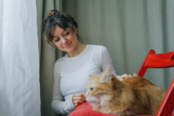 Young woman gently petting her ginger cat relaxing on a red chair next to a window with white...
