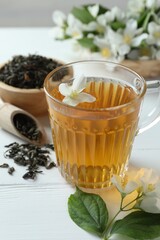 Aromatic jasmine tea in glass cup, flowers and leaves on white wooden table, closeup